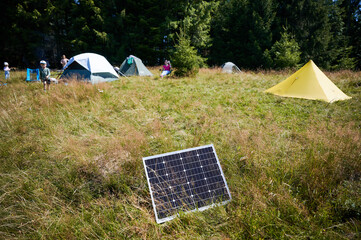 Solar panel in grassy field with tents in background highlights eco-friendly camping setup,...