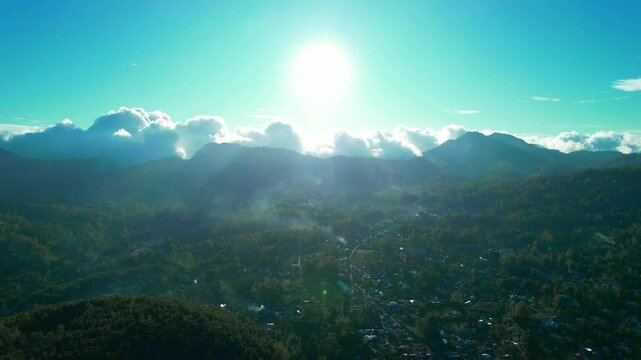 Drone footage showing Bajawa town surrounded by lush green hills and a prominent volcano in the distance under a clear blue sky. Captured during early morning with soft natural light.