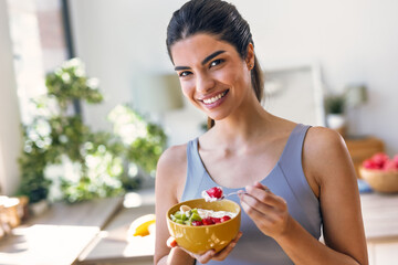 Smiling sporty woman eating a healthy fruit bowl while looking at camera sitting in the kitchen at home