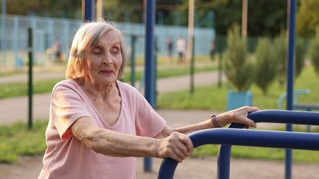 An elderly woman over 80 years old performs a shoulder stretch by bringing her arms in front of her chest and then gently moving them behind her back. This exercise takes place on an outdoor station 