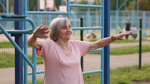 An elderly woman over 80 years old performs a shoulder stretch on an outdoor fitness station. The session takes place during the evening, highlighting active lifestyle.