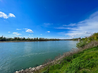  A wide, serene view of the Po River near Cremona, Italy, with lush green banks, distant trees, and a clear blue sky with sparse clouds.