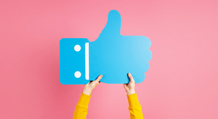 Fototapeta premium Thumbs Up: A person's hands holding a large blue thumbs up sign, against a pink background, communicating endorsement, approval and positive reinforcement.