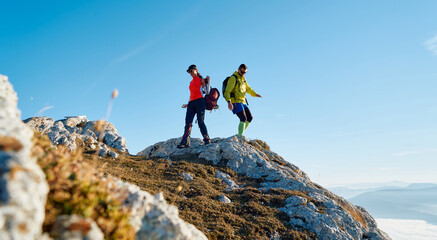 Couple hikers trek up a rocky mountain trail, enjoying a scenic view above the clouds, with majestic peaks in the background.
