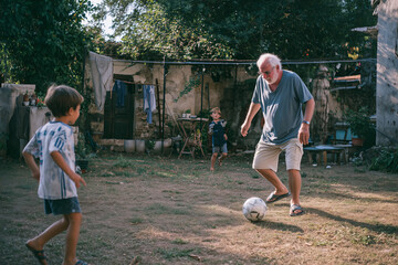 Elderly man playing soccer with children in a backyard garden under sunny skies, creating joyful memories in an outdoor setting