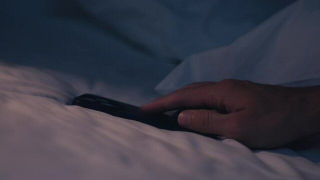 Turning off alarm on your smartphone. Smartphone is displaying snooze button on unmade bed with white sheets, suggesting early morning wake up or difficulty waking up. Smartphone on bed in dim light