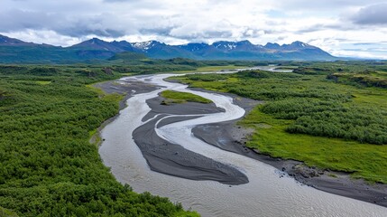 Breathtaking panoramic view of South Iceland's dramatic landscape featu a meande river flowing through lush green vegetation and majestic snow-capped mountains under a
