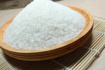 A wooden bowl filled with white crystal sugar placed on a bamboo mat