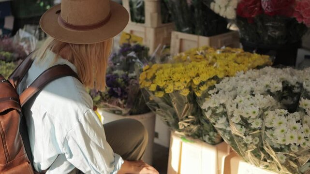 Female traveler wearing sunhat and backpack exploring vibrant flower market, carefully selecting and gently smelling various blossoms, experiencing local culture with relaxed demeanor