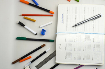 A flat lay of a 2025 calendar planner with a monthly goal page, surrounded by pens, markers, ruler, sharpener, and colored pencils on a white desk.