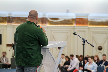 Speaker engages audience in conference room setting with microphone and podium backdrop.