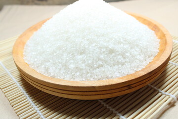 A wooden bowl filled with white crystal sugar placed on a bamboo mat