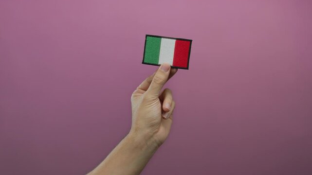 Caucasian man holding an italian flag patch against an isolated pink wall background, showcasing national pride and cultural identity with embroidery details.