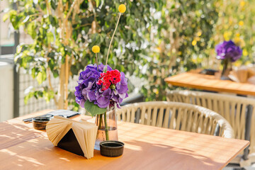 Glass vase with purple hydrangeas, red carnations, and yellow billy balls on an outdoor cafe table in bright sunlight.
