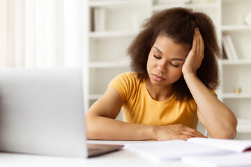 Exhausted young black woman resting her head on hand, eyes closed while sitting at home desk....