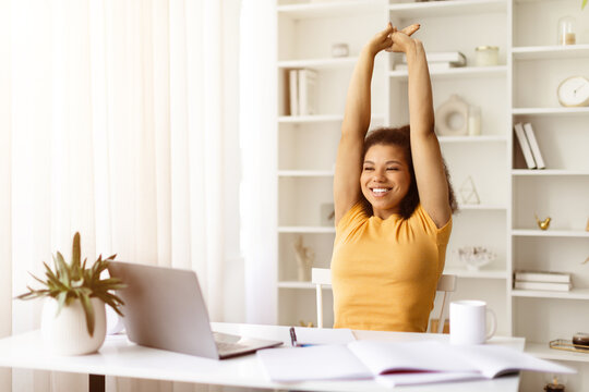 Smiling young black woman stretching arms above head while working from home. Break from study or remote work in a bright and peaceful home office environment, copy space - Powered by Adobe