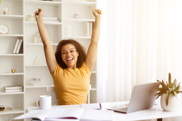 Excited young black woman raising both arms in celebration at home workspace. Achievement, success...