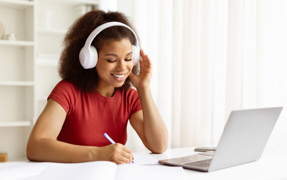 Young black woman writing in notebook and using laptop at home. Focused african american female smiling during online class or remote job from comfortable desk, closeup