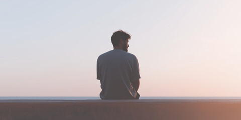 Young man sitting on a concrete ledge, gazing at a serene sunset over the horizon, reflecting on life and enjoying a moment of solitude and tranquility