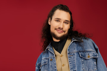 Handsome young man with a beard and long curly hair wearing a denim jacket smiles warmly