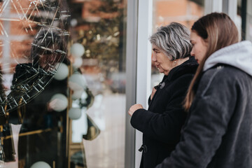 An elderly woman and a young adult admire an intricate item in a glass window display, showcasing a moment of curiosity and connection, with a blurred urban environment framing the scene.