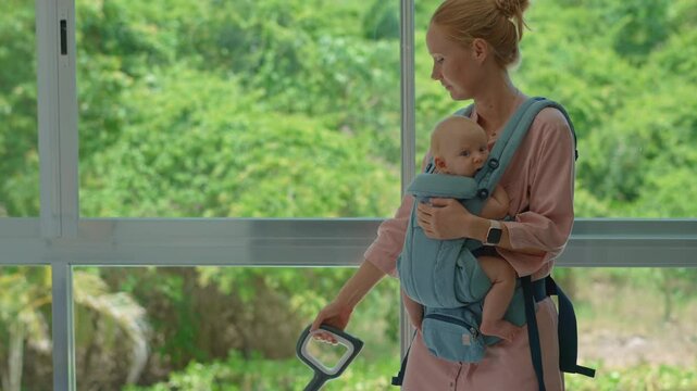 Young mother washes the floor in a bright apartment with a large window and lush greenery outside, while her newborn baby sits safely in a front ergonomic baby carrier