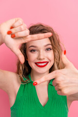 Cheerful young woman in a vibrant green top against a pink background, making a frame with her...