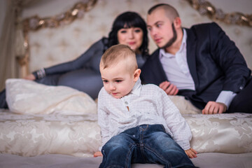 A two year old son sits on the edge of the bed. Dad and pregnant mom are watching him from behind....