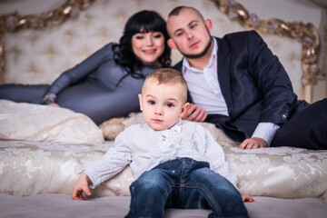 A two year old son sits on the edge of the bed. Dad and pregnant mom are watching him from behind....