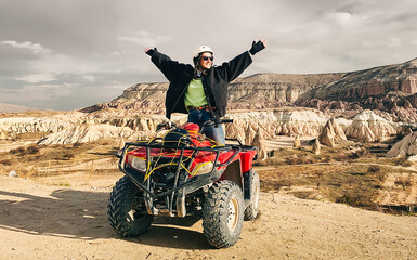 Obraz premium Goreme, Turkey - 26th december, 2024: happy joyful excited woman adventurer hands up sit on red squad over Cappadocia red valley. ATV squad group tour viewpoint. Travel Turkey in winter outdoors