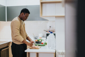 Depicts a man in a well-lit, contemporary kitchen chopping vegetables and preparing food,...