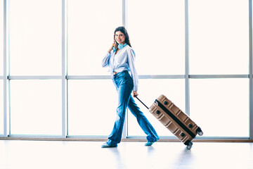 Young woman pulling suitcase at airport terminal, wearing headphones, enjoying her journey,...