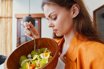 Young woman enjoying a healthy salad in a wooden bowl, wearing a stylish outfit with a warm color...