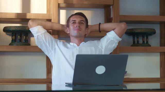Businessman is relaxing at his desk with hands behind his head, enjoying break from work while sitting in front of his laptop in modern office. Stretching after long remote work session