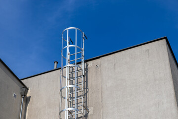 Tall silver industrial caged access ladder firmly attached to a plain grey concrete building wall,...