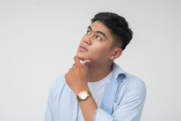 Young Filipino man in light blue long sleeves looks up while resting hand on chin, showing...