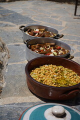 Round clay pot filled with chickpeas mixed with chopped leeks. Two metal pans in the background contain a rich stew with visible chunks of eggplants, tomatoes, and onions (giachni) - Cretan gastronomy