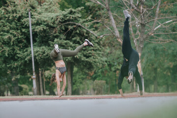 Two people are doing handstands outdoors on a track, wearing athletic clothing, with lush green...