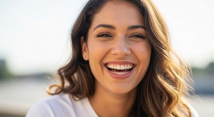 Joyful young woman with perfect white smile and bright eyes laughing in golden sunlight. Close-up portrait capturing genuine happiness and positive energy outdoors.