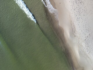 Aerial perspective of sandy coastline with gentle green-tinted ocean waves creating rhythmic...