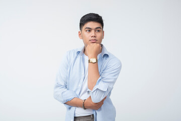 Pensive young Filipino male with chin resting on hand, looking into distance, wearing light blue...