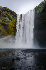 Skogafoss a stunning waterfall in Iceland, tumbles gracefully over cliffs, creating a misty atmosphere. Lush green moss and rocky shores enhance the serene landscape at dusk.