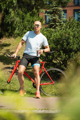 Handsome cyclist in white t-shirt rides standing and drinking water at park. Athlete taking a...