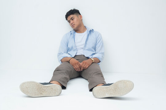 A young Southeast Asian man sits on the floor with slouched posture, head tilted, showing exhaustion and emotional fatigue, lost in thought.