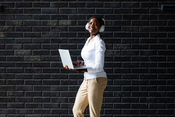Smiling African woman walking by the black brick wall and holding laptop computer.