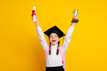 Cheerful schoolgirl celebrating graduation, holding diploma and trophy with excitement against a...