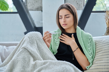 Sad upset teenage girl looking at thermometer sitting at home under blanket