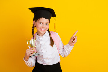 Charming schoolgirl holding a trophy and gesturing on a yellow background wearing a graduation hat...