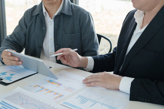 business team gathers around table, using a tablet to discuss economic graphs for trends analyzing ups and downs to strategize their next move together.