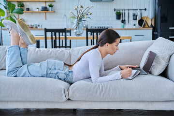 Young student girl study online, lying at home on couch with laptop, notebook